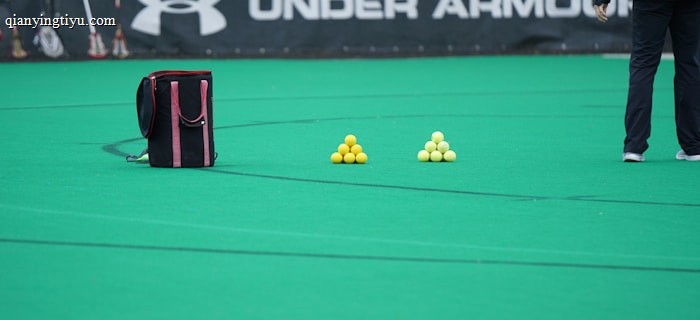 A sports field features a black and pink sports bag, two pyramids of yellow balls, and a portion of a person in dark athletic gear. The background displays a banner with the Under Armour logo.