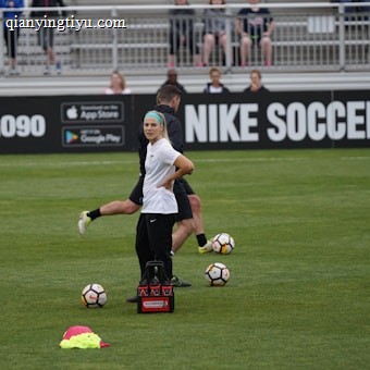 A person in sports attire stands on a soccer field, surrounded by several soccer balls and a carrier with bottles. The field is bordered by advertisements for Nike Soccer and app stores. Several people sit in bleachers in the background.