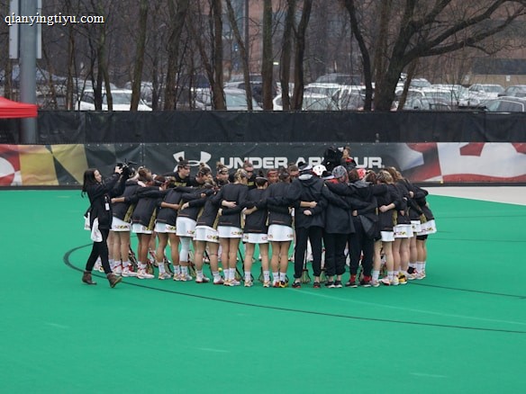 A group of athletes is gathered in a huddle on a sports field, wearing matching uniforms and gear. The ground is green, and there is signage with a brand logo in the background. Trees and parked cars can be seen behind a fence surrounding the field. A person on the left side is capturing the moment with a camera.