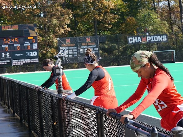 A group of athletes wearing orange uniforms are on the sideline of a sports field, resting against a fence. Behind them, a large digital scoreboard displays team names and scores. The field is bright green, and several banners with sponsor logos and championship wins are visible. The athletes appear to be taking a break, possibly before or during a field hockey game.