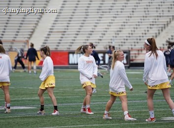A group of young women wearing athletic gear are on a sports field. They are dressed in white jerseys with yellow shorts, featuring a distinctive design at the hem. The field has a green turf surface and the background shows empty bleachers. Some additional figures are visible in the distance, possibly referees or other players.