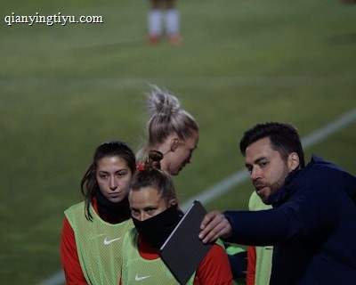 A group of individuals on a sports field are wearing green vests over red attire. One person is holding a tablet and seems to be discussing or showing something to the others. The atmosphere suggests a sports event, likely soccer, indicated by their clothing and the setting. The background is a grassy field.