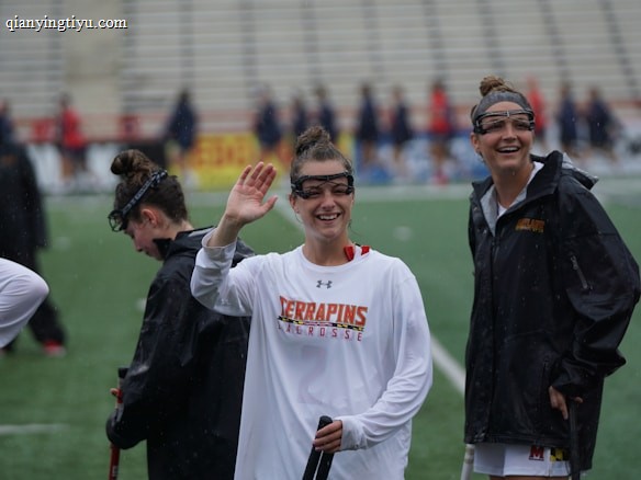 Several athletes in sports attire are on an outdoor sports field, interacting and smiling. One individual in the foreground waves with a friendly expression, while another stands beside her, also smiling. They wear protective goggles and gear associated with lacrosse. The background features indistinct figures in dark clothing against a backdrop of stadium seating.