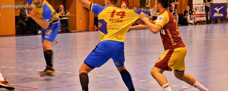 A dynamic indoor sports scene with players in motion on a court. Two athletes in blue and yellow uniforms are engaged in an intense handball game, with one player gripping the other's jersey. The background shows other players, several seated people, and advertisements on the walls.