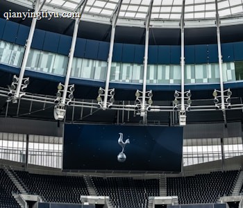 A modern stadium interior featuring a large digital screen with a logo prominently displayed. The seating areas are empty, with dark-colored seats and some structural elements like beams and glass walls visible. A combination of dark blue and neutral tones are prominent.